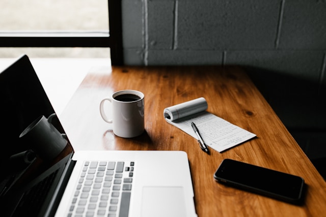 Desk with laptop, mug, notebook, paper, and phone sitting on it.