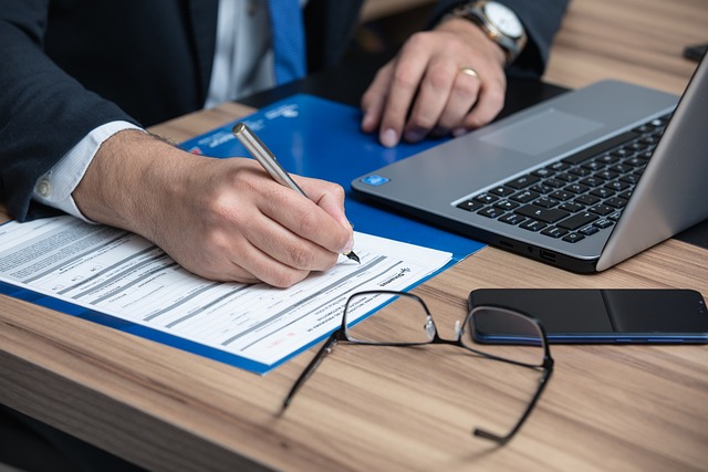 Person writing on documents at a desk with a laptop, phone, and eyeglasses on it.