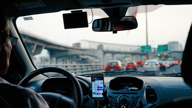 Perspective from the interior of a car looking out through the front windshield. A man with glasses is at the wheel looking out at multiple cars in traffic and using his phone as a navigation system on his dashboard.