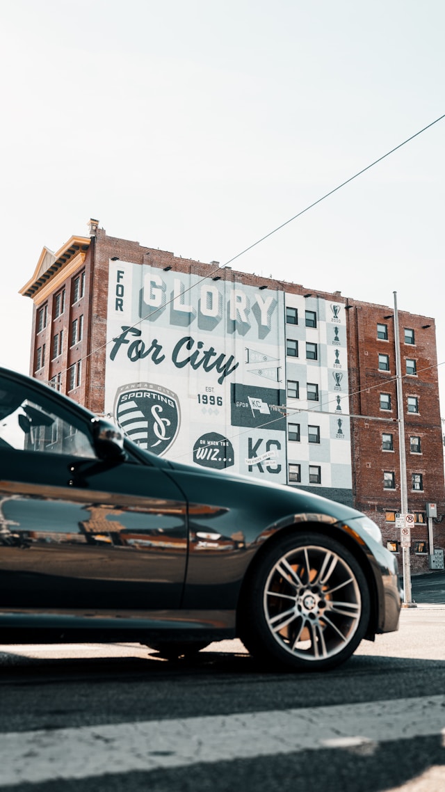 Black car driving on the road in front of a brown building with a mural of Kansas City Sporting KC logo on it.
