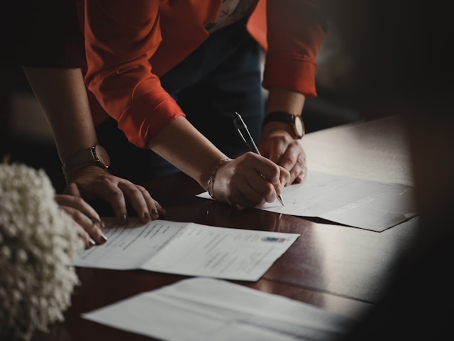 Paperwork on a table being signed by someone wearing a red long sleeve.
