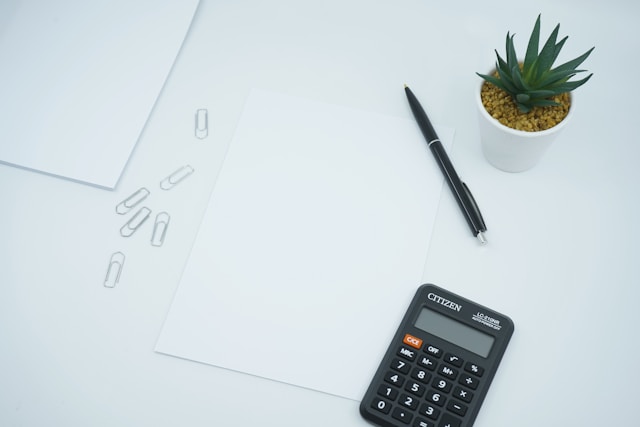 Image of calculator on desk with paper, pen, and plant.