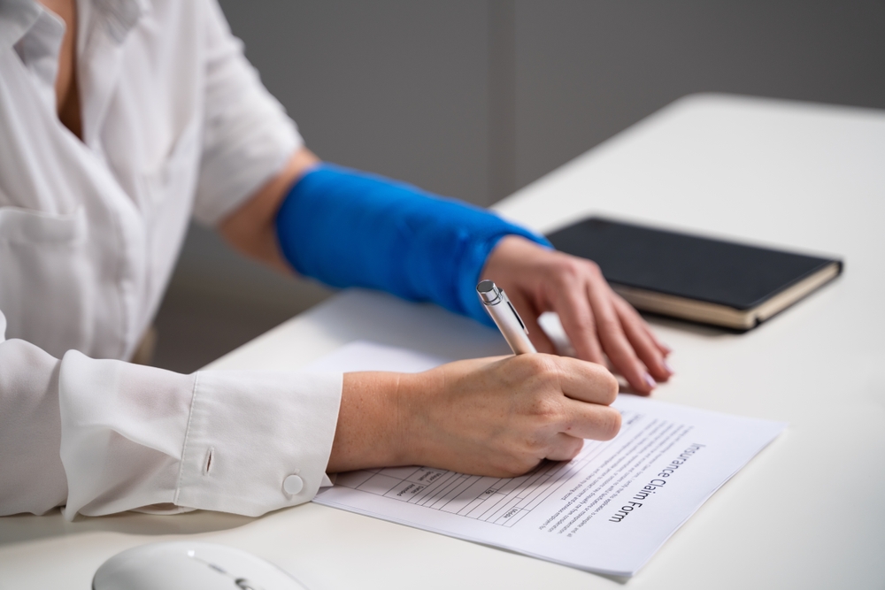 An injured worker with a bandaged arm filling out claim forms at a desk, representing how dual jurisdiction benefits may apply in cross-state workers’ comp claims.