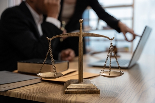 A brass justice scale on a desk in a law office with attorneys discussing a case in the background.