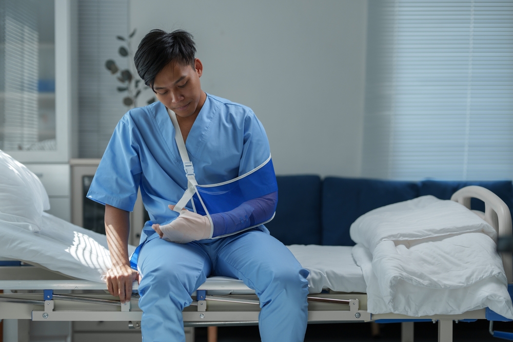 Recovering patient wearing medical scrubs sits on hospital bed, supporting injured arm in blue sling, demonstrating challenges of healthcare and rehabilitation