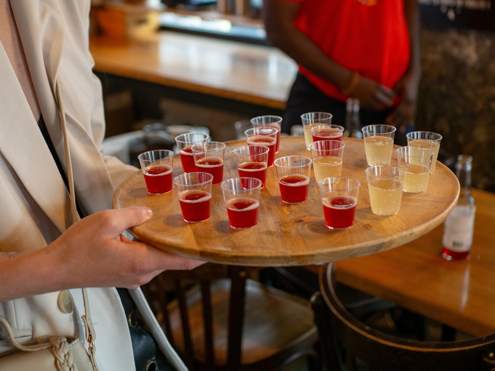 A person holding a wooden tray filled with small cups of red and yellow alcoholic drinks in a bar, illustrating overserving and supporting the topic “Can You Sue a Bar for Overserving.