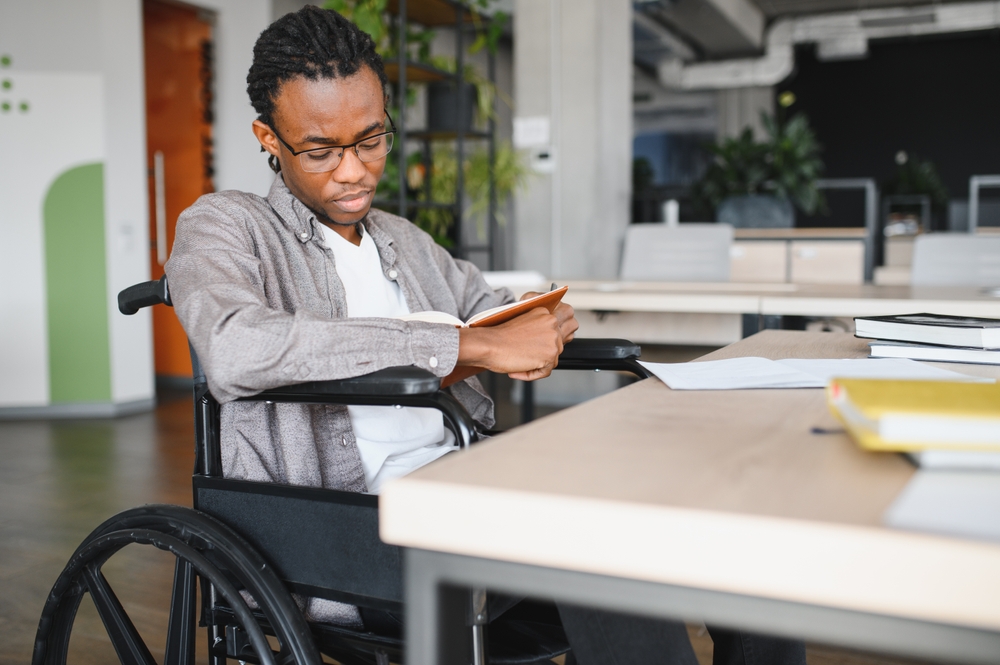 Young black man with disability reading textbook, preparing for exam in accessible college library, embracing inclusive education
